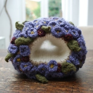 May include: A crocheted flower wreath with purple and green flowers. The wreath is shaped like a circle and has a hole in the center.