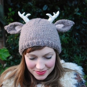 May include: A brown knitted hat with white deer antlers and ears. The hat is worn by a person with brown hair.