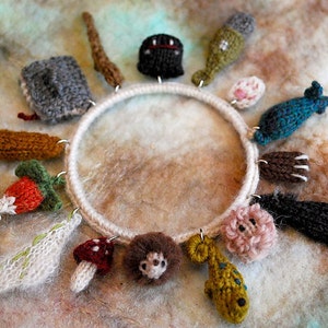 May include: A white circular hoop with various knitted charms hanging from it. The charms include a mushroom, a strawberry, a hedgehog, a fish, a paw print, a leaf, a stick, a bottle, a house, a bird, and a small pink ball.