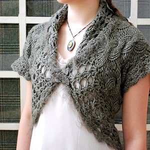 Puede incluir: Una estola de ganchillo gris con un diseño de encaje y un borde festoneado. La estola se lleva sobre una blusa blanca y tiene un cierre de botón decorativo.