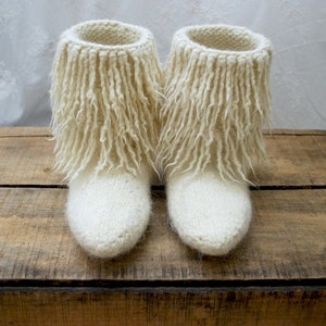 Puede incluir: Un par de botas de punto blancas con flecos largos y esponjosos alrededor de la parte superior. Las botas están sobre una superficie de madera.