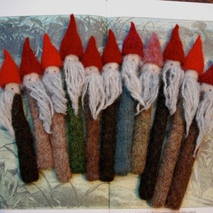May include: A collection of 10 felt gnome ornaments with red hats and white beards. The gnomes are arranged in a row on a vintage book page with illustrations of fairies and mushrooms.