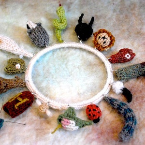 Könnte beinhalten: Ein weißer runder Strickring mit verschiedenen gestrickten Tieranhängern. Die Anhänger umfassen einen Hasen, einen Löwen, einen Kaktus, einen Kolibri, einen Marienkäfer, einen Schmetterling, einen Pilz, eine Socke, einen Fisch und eine Katze.