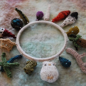 May include: A white knitted ring with various knitted charms attached, including a starfish, a fish, a bird, a mushroom, a branch, and a few other shapes. The charms are all different colours and textures.