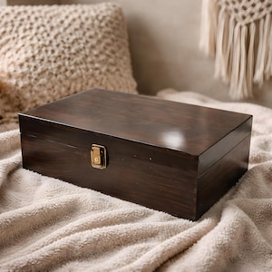 May include: A rectangular, dark brown wooden box with a glossy finish. The box has a brass-colored clasp on the front. The box is resting on a soft, cream-colored blanket.