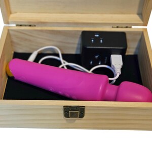May include: A pink vibrator with a yellow tip rests inside an open wooden box lined with black fabric. A black power adapter and a white USB cable are also in the box. The box has a metal clasp.
