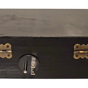 May include: A black wooden box with a rectangular shape. The box features two brass-colored hinges and a latch on the front. The wood grain is visible, and the box appears to be stained or painted black.