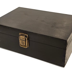 May include: A rectangular black wooden box with a brass-colored clasp. The box has a simple, classic design, with visible wood grain. The clasp is centered on the front, providing a secure closure. The box is likely used for storage or display.