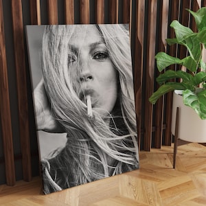Può includere: Stampa in bianco e nero di un ritratto di donna con lunghi capelli biondi, che fuma una sigaretta. L'immagine è presentata su una tela, appoggiata a una parete di legno. Una pianta in vaso è visibile sullo sfondo.