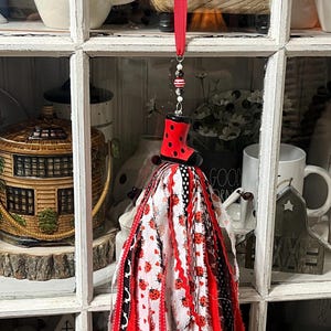May include: A decorative ladybug-themed ornament. The ornament features a red boot with black spots, a beaded strand, and a long, flowing tassel made of red, white, and black fabric strips. The ornament hangs from a red ribbon.