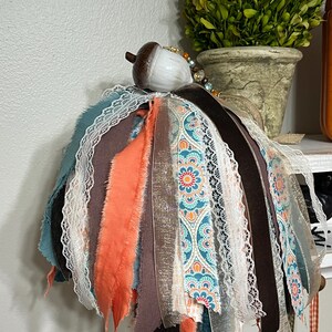 May include: A decorative acorn-topped ornament with a layered design. The ornament features strips of teal, orange, brown, and patterned fabric, accented with white lace. A small, ceramic acorn tops the design, with a beaded accent.