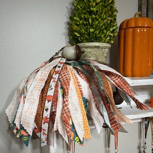 May include: A handmade fall-themed tassel ornament with a wooden acorn top. The tassel is made of various fabric strips in orange, white, and green patterns. The ornament is hanging from a shelf with other fall decorations.