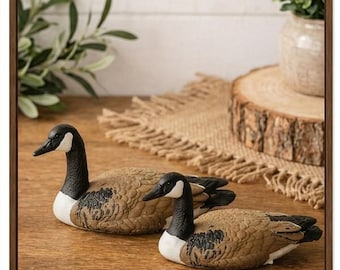 May include: Two decorative goose figurines with black heads, necks, and beaks, and brown bodies with black markings. The geese are resting on a wooden surface. The image is framed in a brown wooden frame.