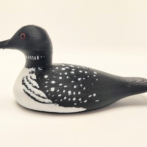 May include: A decorative loon figurine. The bird has a black head and back with white spots and a white belly. The eyes are red, and the beak is black. The figurine is a detailed representation of a loon.