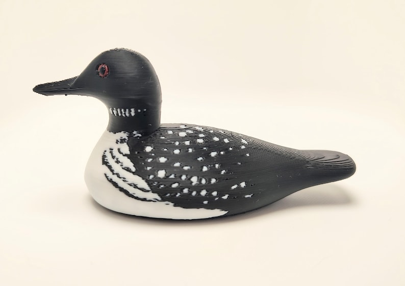May include: A decorative loon figurine. The bird has a black head and back with white spots and a white belly. The eyes are red, and the beak is black. The figurine is a detailed representation of a loon.