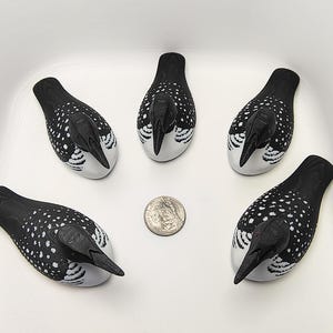 May include: Five decorative loon figurines. Each loon has a black head and back with white spots, a white belly, and a black beak. A U.S. quarter is next to the figurines for size comparison. The loons are arranged in a white container.