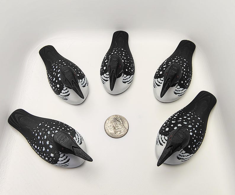 May include: Five decorative loon figurines. Each loon has a black head and back with white spots, a white belly, and a black beak. A U.S. quarter is next to the figurines for size comparison. The loons are arranged in a white container.