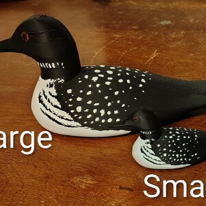 May include: Two wooden loon figurines, one large and one small, are displayed on a wooden surface. The loons are painted black with white speckled patterns on their backs and white bellies. The text "Large" and "Small" are written below each loon.