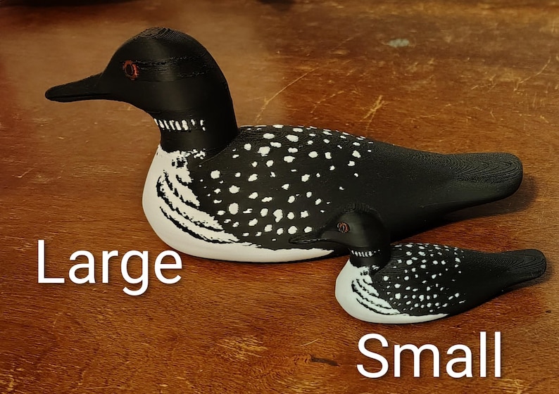 May include: Two wooden loon figurines, one large and one small, are displayed on a wooden surface. The loons are painted black with white speckled patterns on their backs and white bellies. The text "Large" and "Small" are written below each loon.