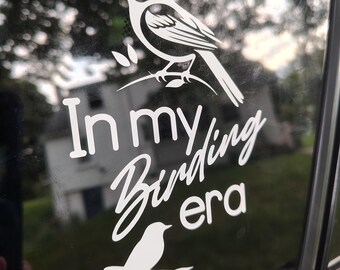 May include: White vinyl decal featuring two bird illustrations and the phrase "In my Birding era." The top bird is perched on a branch, while the bottom bird sits on a branch. The decal is applied to a car window.
