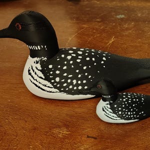 May include: Two decorative loon figurines. The larger loon is black and white with a speckled pattern on its back. The smaller loon mirrors the design. Both have black beaks and red eyes. The figurines are on a wooden surface.