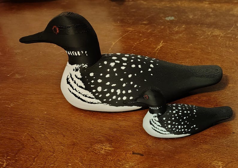 May include: Two decorative loon figurines. The larger loon is black and white with a speckled pattern on its back. The smaller loon mirrors the design. Both have black beaks and red eyes. The figurines are on a wooden surface.
