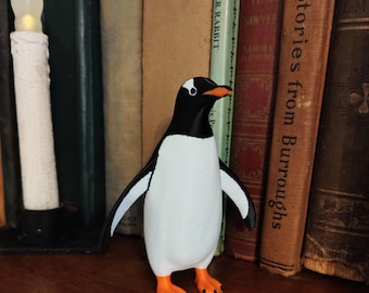 May include: A penguin figurine with a black head and back, white body, and orange feet and beak. The penguin stands on a wooden shelf in front of a collection of vintage books. A white candle is to the left of the penguin.