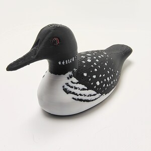 May include: A 3D-printed decorative loon figurine. The bird features a black head and back with white spots, a white belly, and a long, black beak. The loon has red eyes and is resting on a white surface.