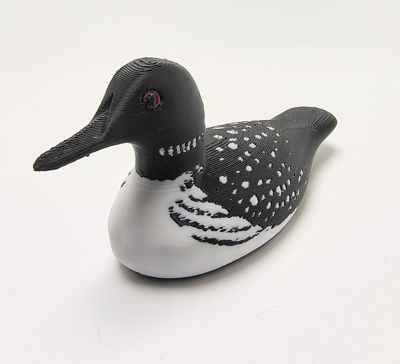 May include: A 3D-printed decorative loon figurine. The bird features a black head and back with white spots, a white belly, and a long, black beak. The loon has red eyes and is resting on a white surface.