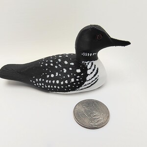 May include: A decorative wooden loon figurine, painted black and white, with a detailed design. The loon has a black head and back, with white spots and a white chest. A quarter is next to the loon for size comparison.