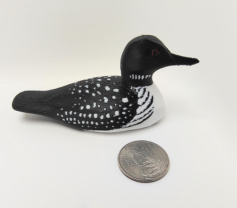 May include: A decorative wooden loon figurine, painted black and white, with a detailed design. The loon has a black head and back, with white spots and a white chest. A quarter is next to the loon for size comparison.