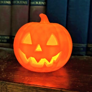 May include: A bright orange, glowing jack-o'-lantern pumpkin lamp with a carved face featuring two triangular eyes, a small triangular nose, and a jagged, toothy grin. The pumpkin has a smooth, rounded surface and a small stem at the top.