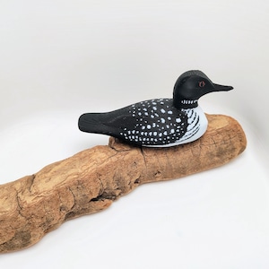 May include: A decorative wooden loon figurine, painted black with white spots, rests on a piece of driftwood. The loon has a black head and beak, with a white and black striped neck. The driftwood is a light brown color.