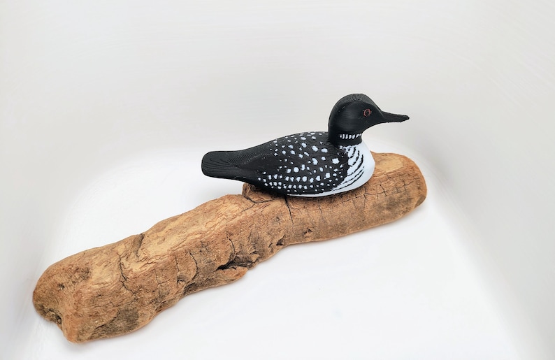May include: A decorative wooden loon figurine, painted black with white spots, rests on a piece of driftwood. The loon has a black head and beak, with a white and black striped neck. The driftwood is a light brown color.