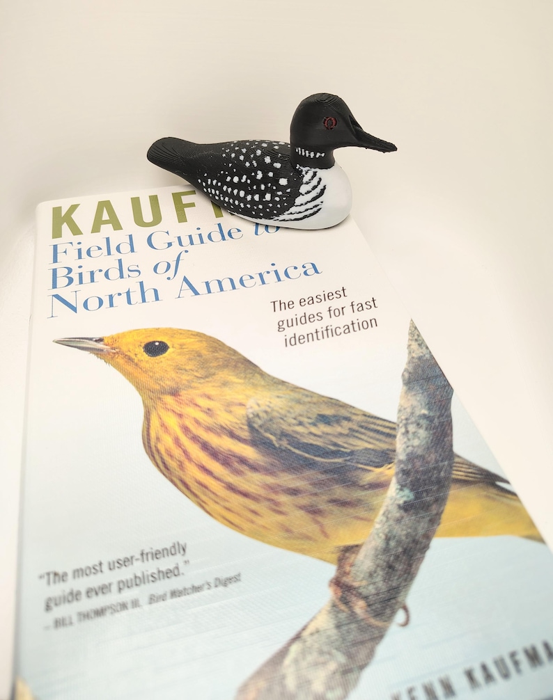 May include: A wooden loon figurine with black, white, and speckled details rests on a field guide titled "KAUFMAN Field Guide to Birds of North America." The book cover features a yellow bird illustration and text that reads "The easiest guides for fast identification."