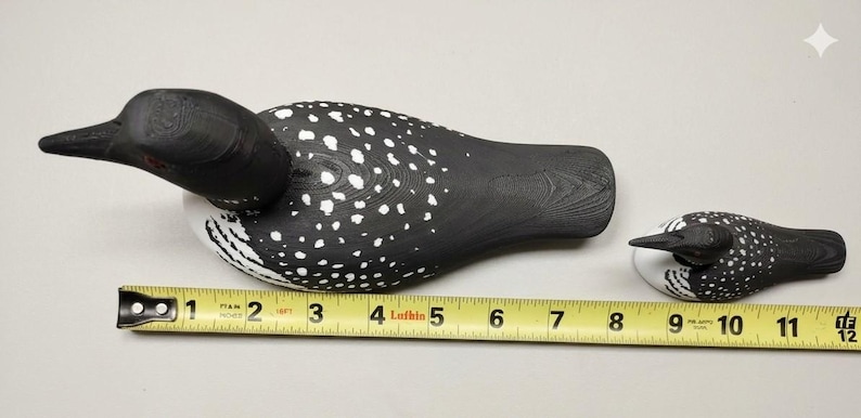 May include: Two wooden loon decoys, one large and one small, are displayed on a white surface. The larger decoy is approximately 10 inches long, and the smaller one is about 4 inches long. Both are painted black with white spots.