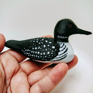 May include: A decorative loon figurine, featuring a black head and back with white polka dots on the wings. The bird has a white belly and a long, black beak. The figurine is held in a hand.