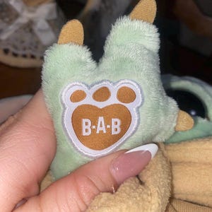 Op de afbeelding: Een pluche knuffelpoot van lichtgroene stof met beige klauwen en een centrale beige voetzool met de witte geborduurde tekst B-A-B. De poot zit vast aan een grotere pluche figuur die gedeeltelijk bedekt is door een beige kledingstuk van zachte stof.