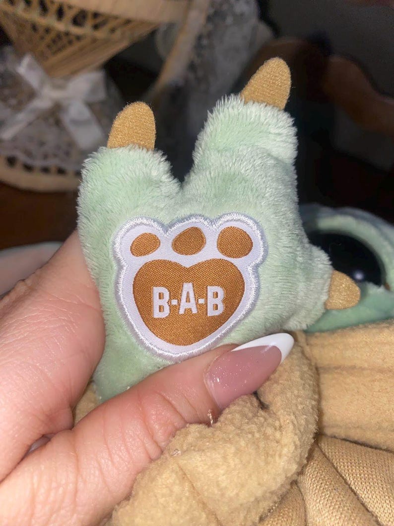 Op de afbeelding: Een pluche knuffelpoot van lichtgroene stof met beige klauwen en een centrale beige voetzool met de witte geborduurde tekst B-A-B. De poot zit vast aan een grotere pluche figuur die gedeeltelijk bedekt is door een beige kledingstuk van zachte stof.