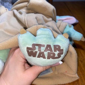 Op de afbeelding: Een knuffel met een lichtgroene voet met drie tenen en het bruine Star Wars-logo op de zool. Het speelgoed draagt een beige, fleece-achtig kledingstuk met capuchon dat het bovenlichaam bedekt, met zachte, lichtgroene ledematen eronder zichtbaar.