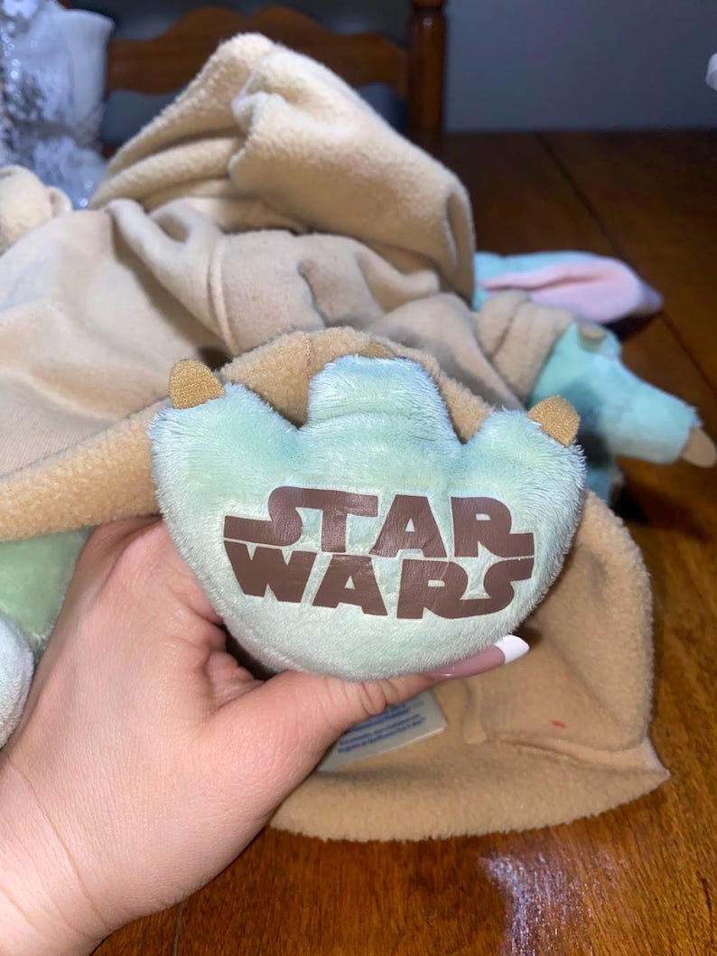 Op de afbeelding: Een knuffel met een lichtgroene voet met drie tenen en het bruine Star Wars-logo op de zool. Het speelgoed draagt een beige, fleece-achtig kledingstuk met capuchon dat het bovenlichaam bedekt, met zachte, lichtgroene ledematen eronder zichtbaar.