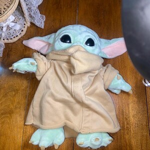 Op de afbeelding: Een knuffel van een lichtgroen wezen met grote zwarte ogen, puntige oren met roze details aan de binnenkant en kleine klauwtjes aan de handen en voeten. Het wezen draagt een beige fleece gewaad met een hoge kraag en lange mouwen.