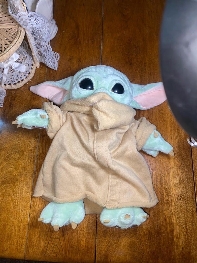 Op de afbeelding: Een knuffel van een lichtgroen wezen met grote zwarte ogen, puntige oren met roze details aan de binnenkant en kleine klauwtjes aan de handen en voeten. Het wezen draagt een beige fleece gewaad met een hoge kraag en lange mouwen.