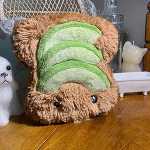 May include: A plush toy shaped like a piece of toast with avocado. The toast is brown with a shaggy texture, and the avocado is green with a ribbed design. Two black button eyes are visible.