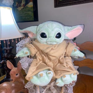 Op de afbeelding: Een knuffel van een groen wezen met grote zwarte ogen, puntige oren en roze details aan de binnenkant van de oren. Het wezen draagt een beige fleece gewaad en heeft kleine klauwen aan de handen en voeten. Het woord STAR is zichtbaar op &eacute;&eacute;n voet.