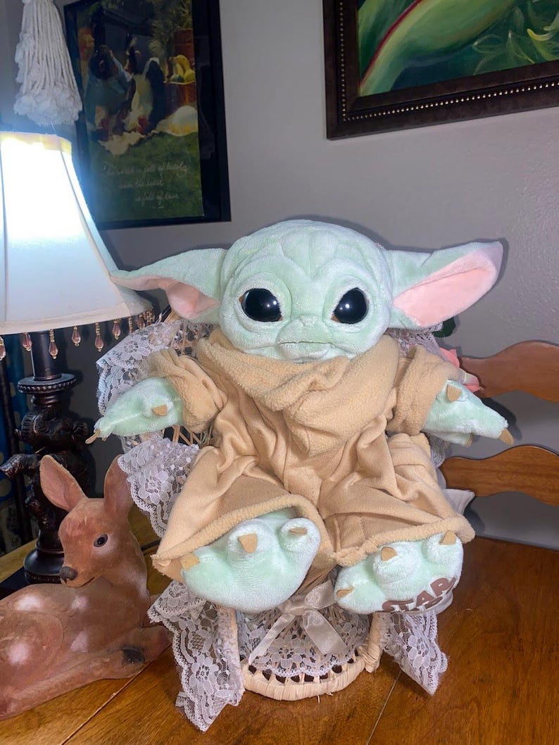 Op de afbeelding: Een knuffel van een groen wezen met grote zwarte ogen, puntige oren en roze details aan de binnenkant van de oren. Het wezen draagt een beige fleece gewaad en heeft kleine klauwen aan de handen en voeten. Het woord STAR is zichtbaar op &eacute;&eacute;n voet.