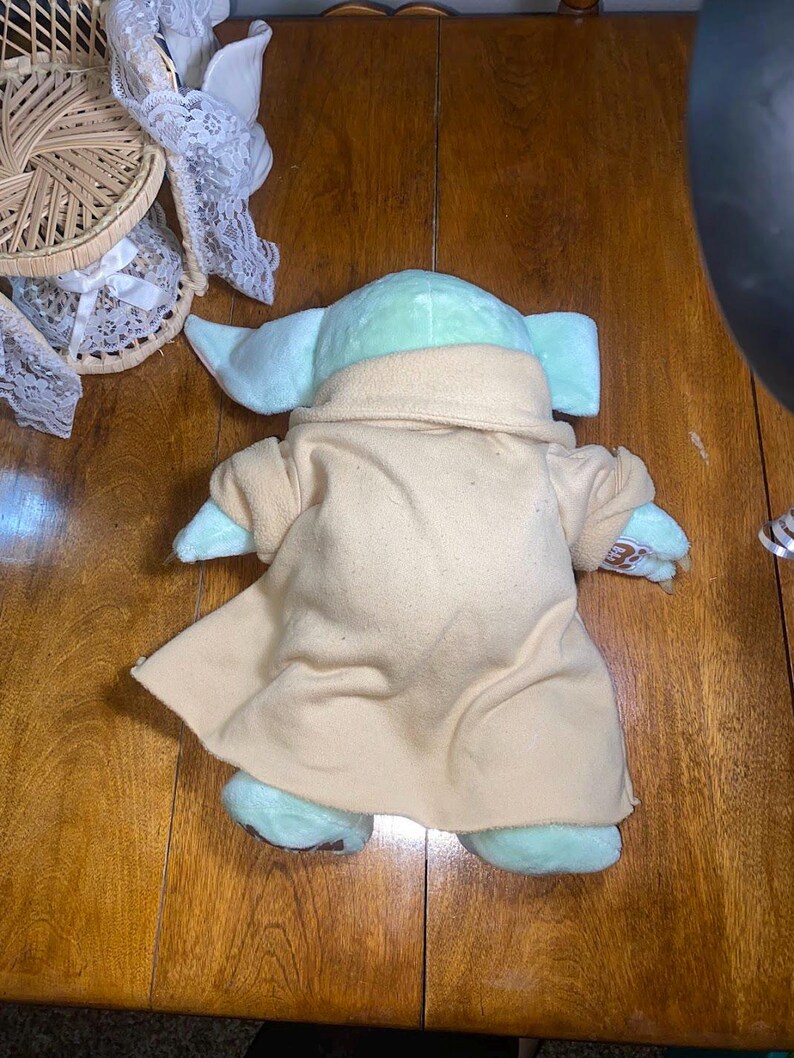 Op de afbeelding: Een lichtgroene knuffel met grote oren, gekleed in een beige gewaad met een brede kraag. De knuffel heeft een Build-A-Bear Workshop logo-patch op de linkerpoot. Het personage wordt van achteren getoond, inclusief de volledige lengte van het gewaad.