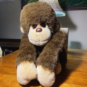 May include: A plush brown monkey with a light tan face, hands, and feet. The stuffed animal has a friendly expression with a brown nose and dark eyes. The monkey is sitting on a wooden surface.