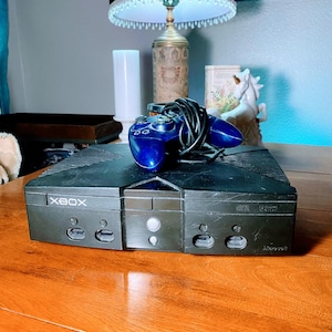 May include: A black Xbox console with a blue controller and a black cord. The console has the word "XBOX" printed on the front. The controller is resting on top of the console. The console is sitting on a wooden table.