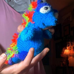 May include: A handmade blue crochet dragon with a rainbow mane and tail. The dragon has a white eye and a smiling face. The toy is held in a person's hands.